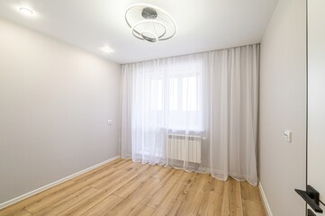 bright, empty room with wooden floors and light gray walls. A window with sheer white curtains provides natural light. A modern ceiling light fixture is installed