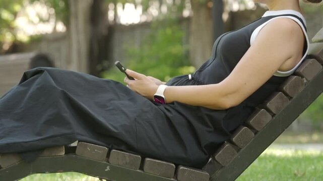 A woman lies back on a wooden lounge chair, focused on her phone. She enjoys a peaceful moment surrounded by greenery in a sunny park in Simferopol, Crimea.