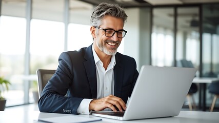 Smiling middle aged businessman with gray hair and beard wearing glasses and a suit works on a laptop in a modern office