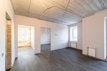 An empty, unfinished room with a concrete ceiling, dark wood floors, white walls, and exposed brick. Radiators are under the windows