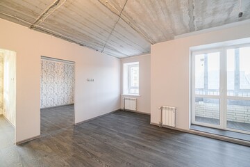 Empty room with dark wood flooring, light pink walls, a bare concrete ceiling, and white windows. One doorway leads to another wallpapered room