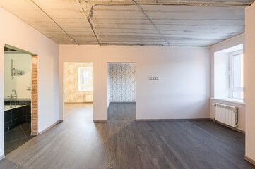 bright, unfinished room with dark wood floors, bare concrete ceiling, and multiple doorways. One doorway leads to a bathroom, others to wallpapered rooms. A window with a radiator is present