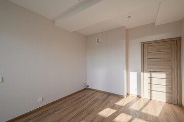 An empty room with light beige walls, wooden floor, and a light-colored door. Sunlight streams in,...