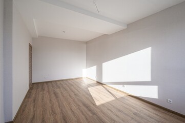 An empty room with wood floors and white walls. Sunlight streams in, creating bright shapes on the wall. A door is visible on the left side