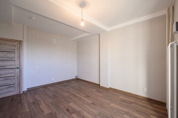 Empty room with wooden floor, light walls, and a door on the left. A simple lightbulb hangs from the white ceiling