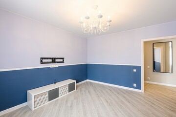 bright, empty room with two-tone walls (blue and lavender), light wood floors, a white shelf unit, and a white chandelier. A doorway leads to another room with a mirror
