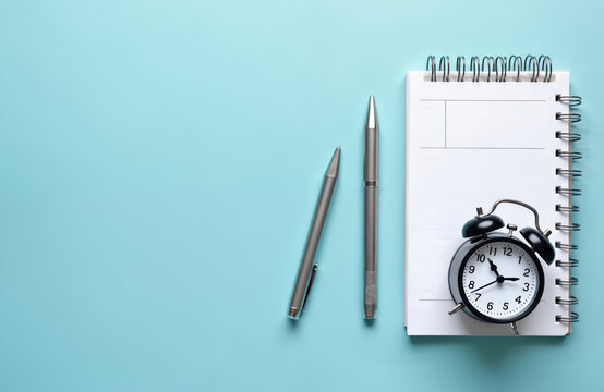 Spiral notebook with blank page on blue background. Silver pen lies beside. Alarm clock shows time, ready to wake. Productive scene, organized desk, daily tasks, schedule, planner, diary, agenda,