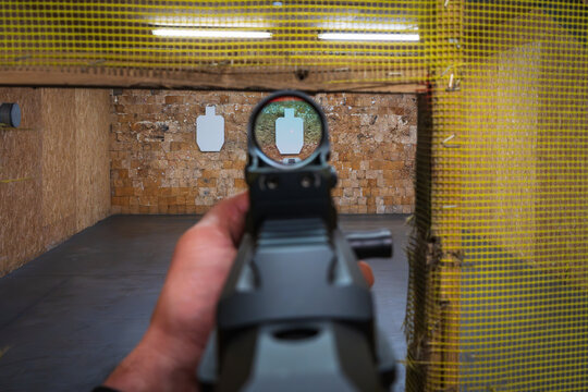 First-person view: a shooter aims a modern carbine with a red dot sight at targets at an indoor shooting range for marksmanship training.