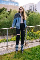Smiling young woman with inline skates enjoying a sunny afternoon in a city park, leaning on a fence