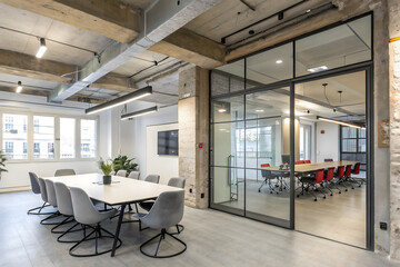Modern coworking office, featuring a spacious meeting room with a large table, gray chairs, exposed concrete beams, and a transparent glass partition