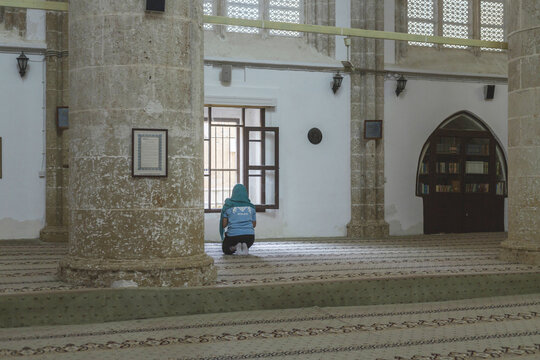 Varosha, Cyprus - 25 June 2025: View of a solitary figure kneeling in prayer inside a weathered, stone-columned mosque, light filtering through barred windows onto patterned carpets.