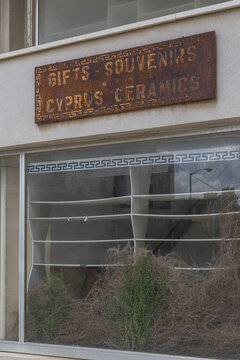 Varosha, Cyprus - 25 June 2025: View of an abandoned storefront with a rusty "Gifts Souvenirs Cyprus Ceramics" sign, overgrown with dried plants behind the dusty window.