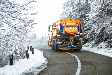 Snowplow truck spreading salt on a winding mountain road during heavy winter conditions. Public services in action for safe driving.