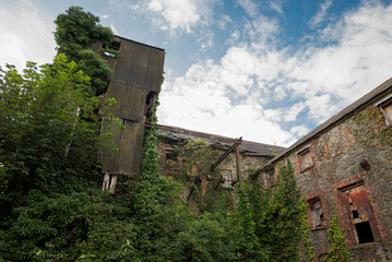Ruins of an abandoned, industrial building shows crumbling stone and brick, with boarded-up windows and ivy climbing the surface. Photo taken in Inniskeen, County Monaghan, Ireland