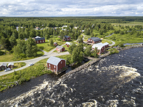 Aerial view of red wooden buildings nestled along the Torne River's rushing waters, a vibrant contrast against the lush green landscape, Haparanda, Norrbotten County, Sweden.