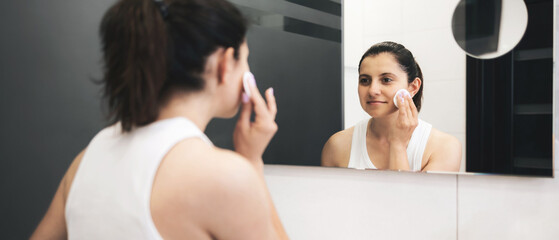 Young woman in bathroom cleansing her face with a cotton pad and facial toner, removing makeup in front of a mirror, facial skincare routine