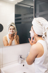 Young woman cleansing and scrubbing her face with cosmetics in the bathroom in front of a mirror, facial skincare routine