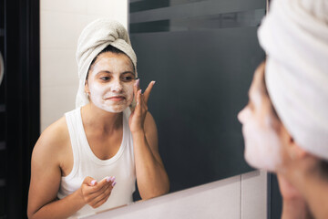 Young woman cleansing and scrubbing her face with cosmetics in the bathroom in front of a mirror, facial skincare routine