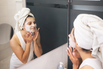 Young woman cleansing and scrubbing her face with cosmetics in the bathroom in front of a mirror, facial skincare routine