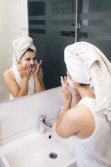 Young woman cleansing and scrubbing her face with cosmetics in the bathroom in front of a mirror, facial skincare routine
