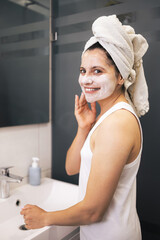 Young woman cleansing and scrubbing her face with cosmetics in the bathroom in front of a mirror, facial skincare routine