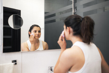 Young woman in bathroom cleansing her face with a cotton pad and facial toner, removing makeup in front of a mirror, facial skincare routine