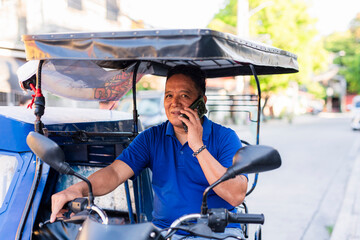 Focused Southeast Asian tricycle driver in blue shirt holds a phone to his ear while looking...