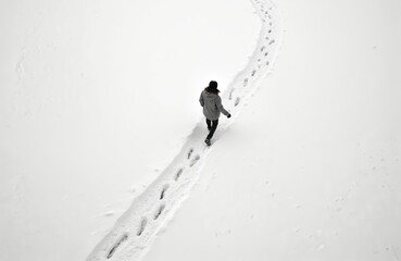 Woman walks through snowy landscape. Gray jacket and black pants. Aerial view of snowy field with winding path. Footprints on trail. Fresh winter activity outdoors.