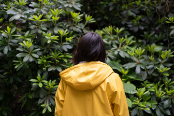 Woman in yellow raincoat with her back to the camera, facing green foliage. Concept: peaceful...
