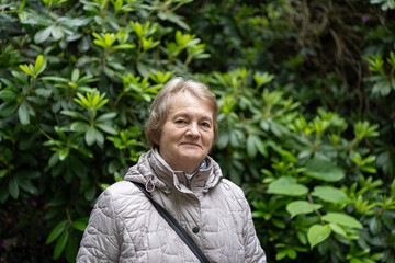Smiling elderly woman in a jacket standing before lush green foliage. Concept: joyful aging, nature...