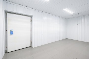 An empty, brightly lit cold storage room with white corrugated metal walls, a white door with a...