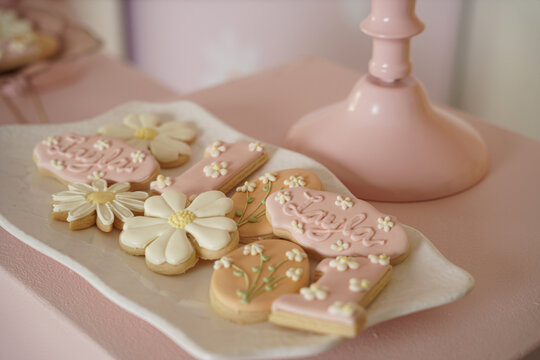Custom sugar cookies decorated with pastel pink icing, daisy flowers, and "Layla" lettering. Includes number one shapes&mdash;ideal for baby girl birthdays, spring parties, and dessert tables.