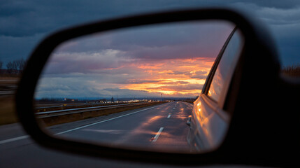 Car side mirror reflecting an open highway and dramatic sunset sky with glowing clouds, creating a scenic and nostalgic road trip moment