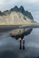 Two adventurers stand on the wet black sands of Stokksnes beach, gazing at the majestic Vestrahorn...