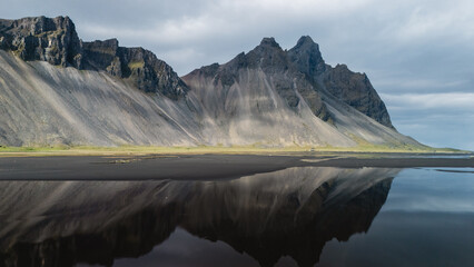 Majestic Vestrahorn mountain rises sharply against a dramatic sky, mirrored in calm waters at...