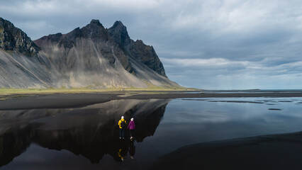 Two hikers stand on the black sand beach at Stokksnes, admiring the breathtaking reflection of...