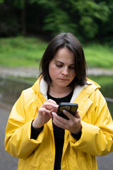 Vertical shot of woman in yellow raincoat scrolling phone in forest. Concept: digital connection in nature, modern exploration, solitude and signal.