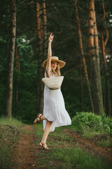 Happy tourist wearing white dress and straw hat is dancing in the forest