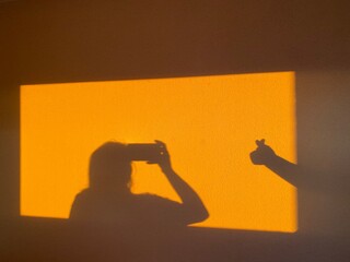 Female Silhouette Taking Photo with Smartphone and Hand Heart Gesture on Golden Sunlit Wall,...