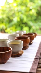 Several small teacups, some wooden, some ceramic, filled with light-colored liquid, on a mat, outdoor setting