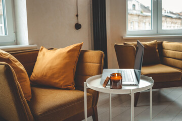 Cozy home interior with mustard armchair, burning candle in glass jar, and laptop on a small white...
