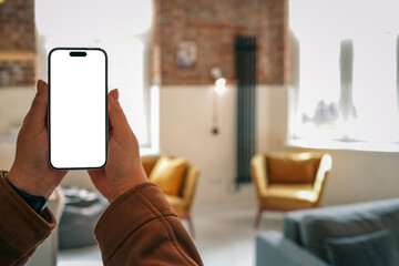 Person holding smartphone with blank screen in cozy loft interior with mustard armchairs, gray...