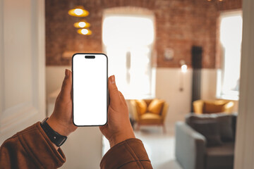 Person holding smartphone with blank screen in cozy loft interior with mustard armchairs, gray...