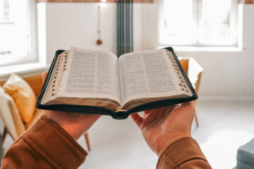 Person holding an open Bible in cozy modern living room with soft natural light. Faith, reading,...