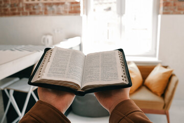 Person holding an open Bible in cozy modern living room with soft natural light. Faith, reading,...