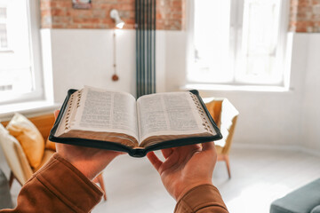 Person holding an open Bible in cozy modern living room with soft natural light. Faith, reading,...