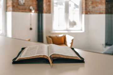 Open Bible resting on table in sunlit modern loft interior with cozy armchairs and warm atmosphere....