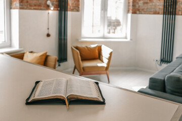 Open Bible resting on table in sunlit modern loft interior with cozy armchairs and warm atmosphere....