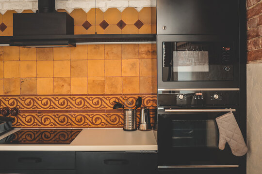 Modern kitchen with black appliances, beige and terracotta tiles, and vintage ceramic pattern. Oven with mitt, utensils, and coffee pot on clean countertop
