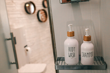 Minimalist bathroom shelf with labeled body wash and shampoo dispensers in white ceramic bottles with wooden pumps, modern toilet visible in background